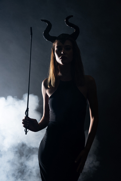 sylwetka młodej kobiety z rogami trzymającej chłostę na czarno z dymem  - Zdjęcie, obraz