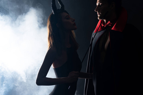handsome man in cloak looking at girl with horns holding flogging whip on black with smoke  - Photo, Image