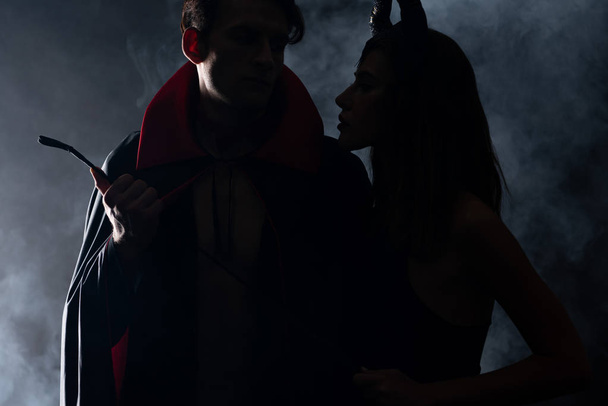 silhouette of man holding flogging whip near woman on black with smoke  - Foto, Bild