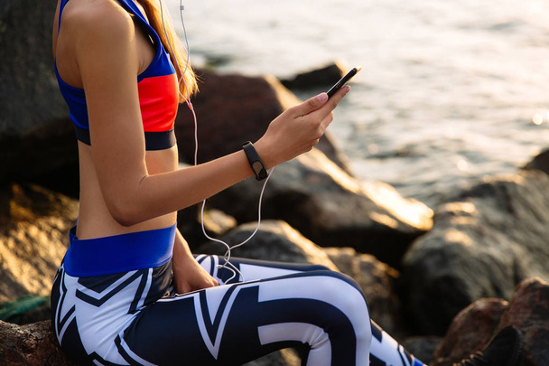 Sportive woman using a smartphone, sitting near the ocean - Фото, зображення
