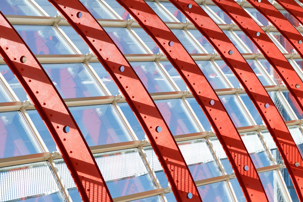 Okenní systémy drží zakřivené červené kovové konstrukce. Sklem proniká jasné sluneční světlo. Ilustrace pevnosti a vzdušnosti železné konstrukce. - Fotografie, Obrázek