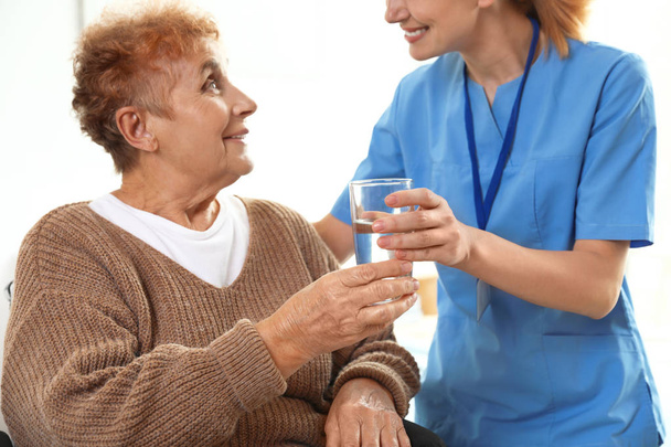 屋内の高齢女性に水のグラスを与える看護師。医療援助 - 写真・画像