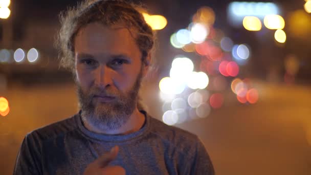 Mosolygó jóképű fehér szakállas, hosszú hajú fiatalember egyenesen a kamerába néz, és azt mutatja hüvelykujját fel a háttérben mozgó esti fények egy város. - Felvétel, videó