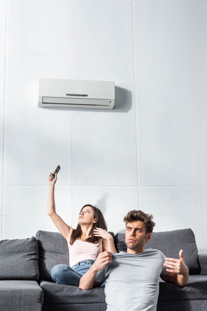 attractive girlfriend switching on air conditioner and handsome boyfriend looking away  - Фото, изображение