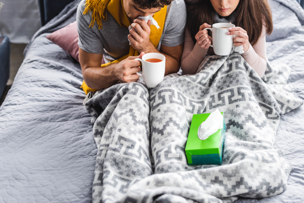 cropped view of sick girlfriend and boyfriend sneezing and holding cups of tea  - Фото, зображення
