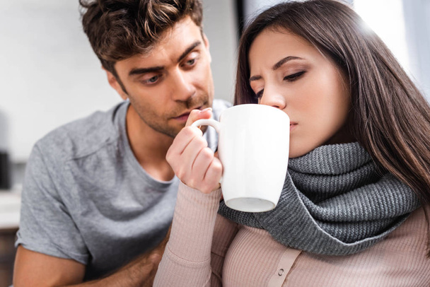 aantrekkelijke en zieke vrouw drinken thee en man kijken naar haar  - Foto, afbeelding