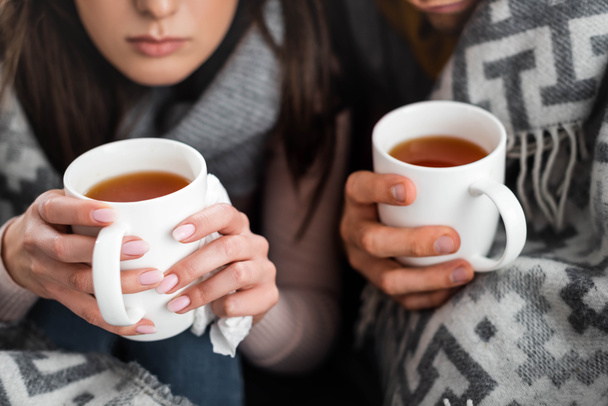 cropped view of sick girlfriend and boyfriend covered with blanket holding cups of tea  - Foto, imagen