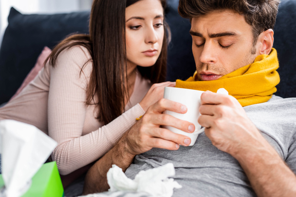 girlfriend looking at sick boyfriend in scarf with cup of tea  - Fotó, kép