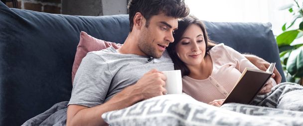 panoramic shot of girlfriend reading book and boyfriend holding cup of tea  - Фото, изображение
