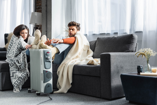 attractive girlfriend and boyfriend covered with blankest warming up near heater  - Φωτογραφία, εικόνα