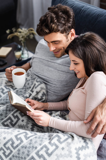 smiling girlfriend reading book and boyfriend holding cup of tea  - Fotografie, Obrázek