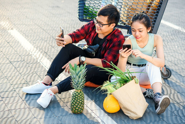 Attractive vietnamese boy and beauiful girl sitting on the parking near shopping mall and uses their smartphones. - Foto, Bild