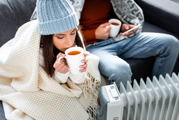 selective focus of attractive girlfriend with cup of tea warming up near heater  - Photo, Image