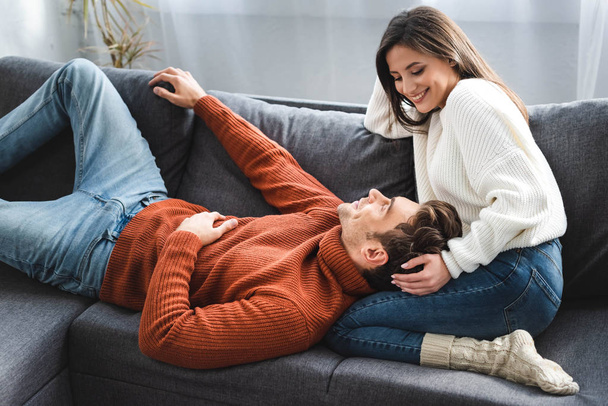 přítel leží na kolenou atraktivní přítelkyně ve svetru a usmívá se v bytě  - Fotografie, Obrázek