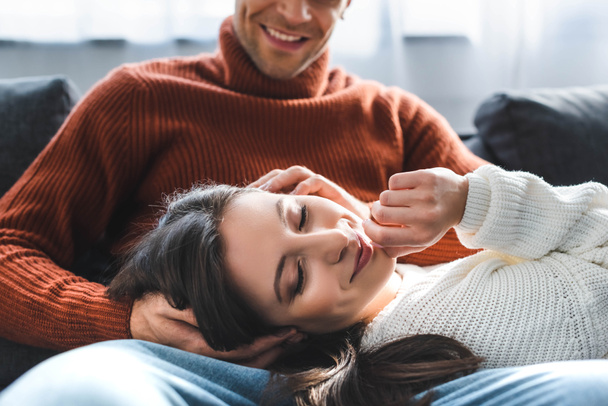 selektivní zaměření atraktivní přítelkyně ve svetru s úsměvem a ležící na kolenou přítele  - Fotografie, Obrázek