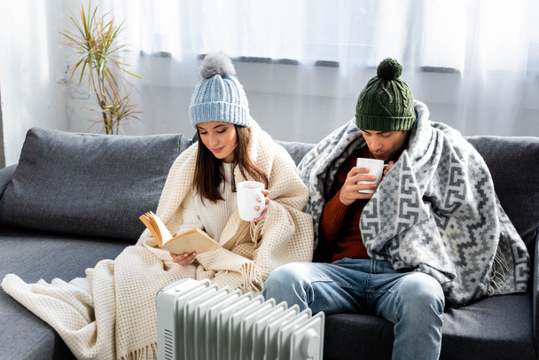 attractive girlfriend reading book and boyfriend in winter outfit with cup warming up near heater  - Photo, Image