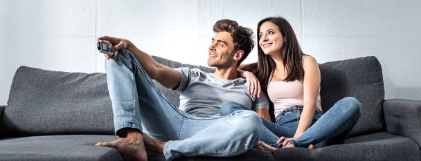 panoramic shot of attractive girlfriend and handsome boyfriend with remote controller sitting on sofa  - Foto, Imagen