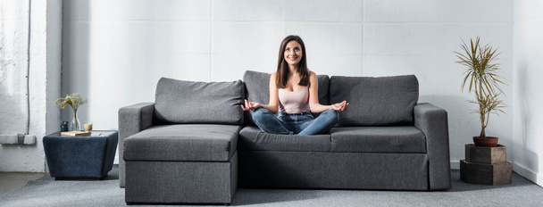 Panoramaaufnahme einer attraktiven und lächelnden Frau in Lotus-Pose, die auf einem Sofa sitzt  - Foto, Bild