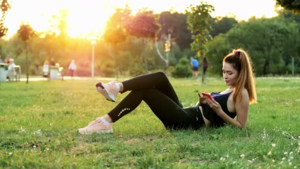 Sportowy Cute Kobieta używać telefonu komórkowego, leżąc na trawie w parku miejskim na zachodzie słońca, mają przerwę w czasie treningu - Materiał filmowy, wideo