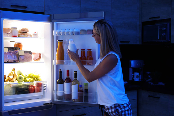 Woman taking bottle with milk out of refrigerator in kitchen at night - Foto, Imagem