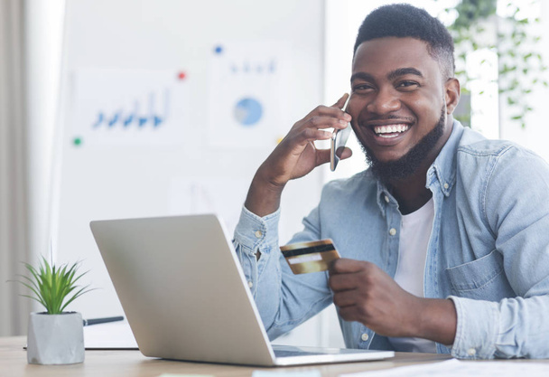 Cheerful black guy talking on phone and holding credit card - Fotó, kép