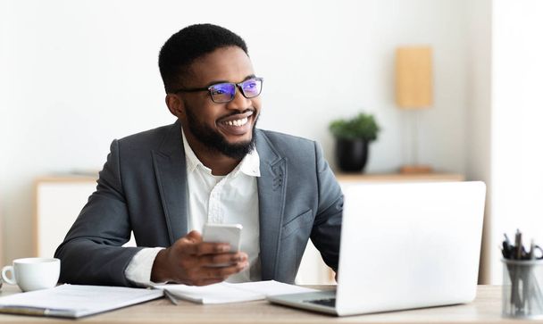 Portrait of smiling black businessman sitting at workplace with cellphone - Zdjęcie, obraz