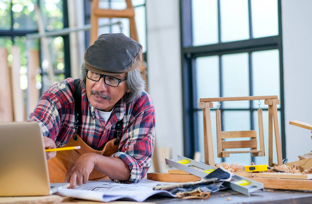 Stary azjatycki rzemieślnik działa jak praca z laptopem o swojej działalności i siedzieć w miejscu pracy. - Zdjęcie, obraz
