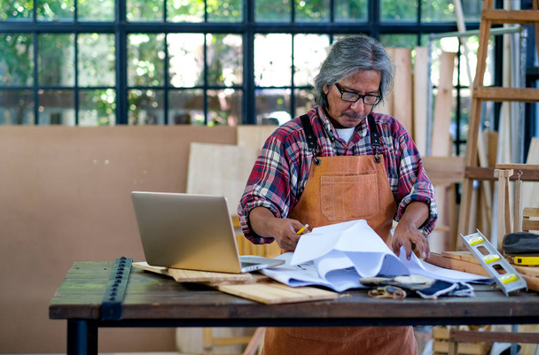 Starý asijský řemeslník stojí před laptopem a přemýšlí o svém procesu práce s papírem a nějakými nástroji na pracovišti během dne. - Fotografie, Obrázek