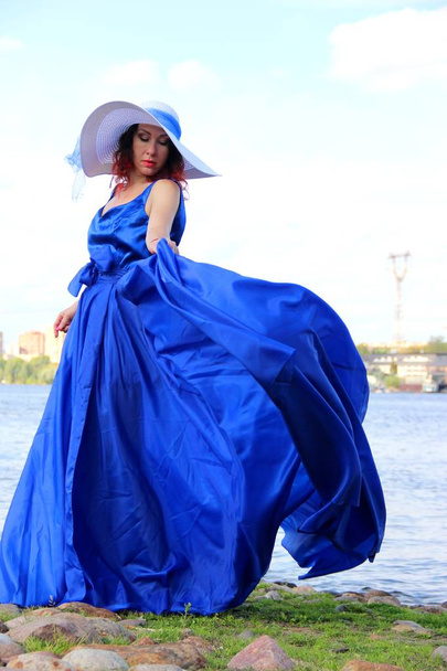 豪華なロングブルーのドレスを着た女性川の岸や風の強い夏の日に - 写真・画像