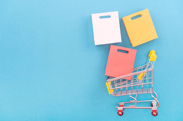 Abstract design element, concept of annual sale, shopping season - mini yellow cart with paper bag isolated on pastel blue background, top view, flat lay. - 写真・画像