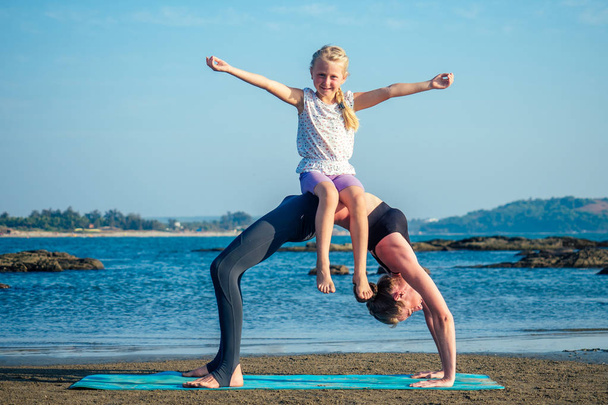 beautiful and flexible mom and charming daughter practicing yoga and doing stretching exercises on the beach - Photo, Image