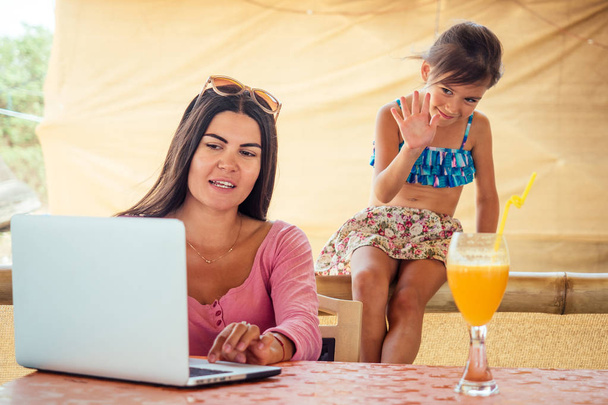 beautiful freelancer-mom and her cute daughter call on video communication with a laptop in a cafe on vacation - Foto, imagen