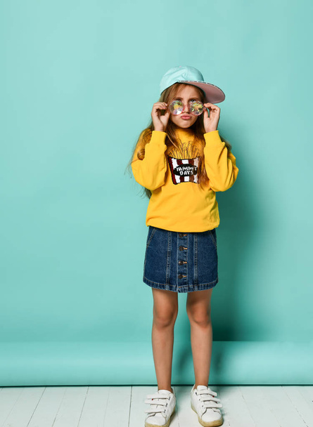 Pretty fashionable young teenager with a lovely smile posing in a baseball cap and yellow hoodie on a blue background - Foto, imagen