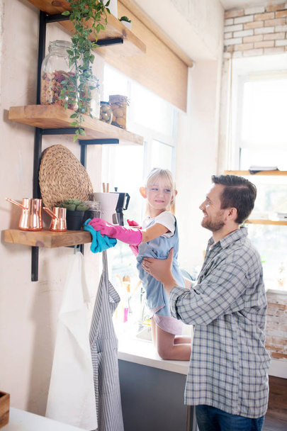 Smiling dad lifting daughter while dusting shelves - Foto, Imagen