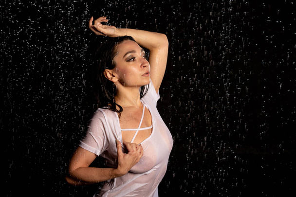 Young expressive wet sexy woman wearing white t-shirt and shorts on the dark background with droplets, water studio - Foto, imagen