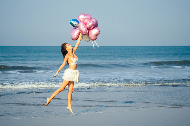 νεαρό και όμορφο κορίτσι που κρατάει μπαλόνια ηλίου αέρα, τρέχει και πηδάει από χαρά στη θάλασσα. έννοια διακοπών και διακοπών - Φωτογραφία, εικόνα