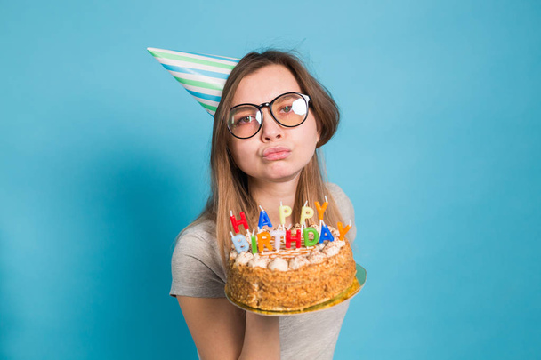 Charming merry crazy young girl student in congratulatory paper hat holding a happy birthday cake in her hands standing against a blue background. Advertising space. - Fotoğraf, Görsel