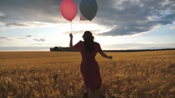 Rear view of happy girl in red dress running through golden wheat field with balloons in hand at sunset. Young woman with brown hair having fun while jogging among barley meadow. Freedom concept - Footage, Video
