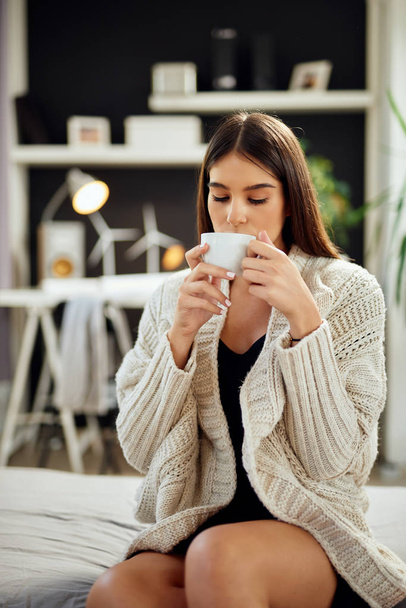 Кавказька жінка, одягнена в бежевий светр, сидить на ліжку і тримає свіжу ранкову каву.  - Фото, зображення