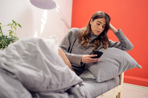 Kaukasisch brunette met bril en gekleed in pullover zitten op het bed in de ochtend en met behulp van smart phone. - Foto, afbeelding