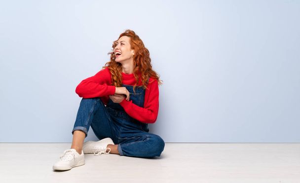 Redhead woman with overalls sitting on the floor happy and smiling - Фото, изображение