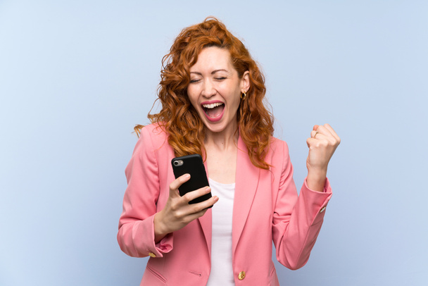 Redhead woman in suit over isolated blue wall with phone in victory position - Photo, Image