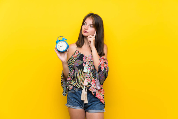 Caucasian girl in colorful dress over isolated yellow background holding vintage alarm clock - Photo, Image