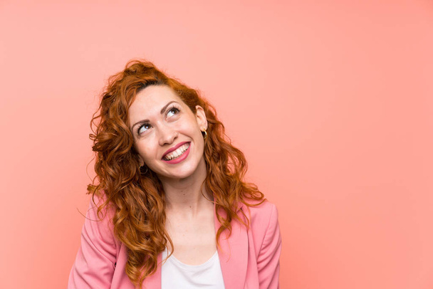 Redhead woman in suit over isolated pink wall laughing and looking up - Foto, afbeelding