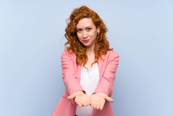 Redhead woman in suit over isolated blue wall holding copyspace imaginary on the palm to insert an ad - Photo, Image