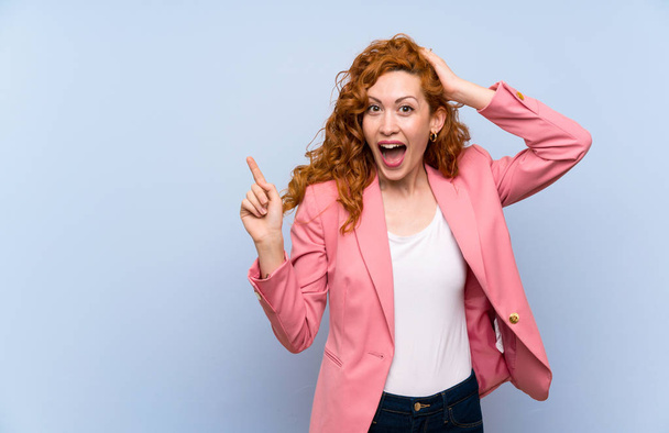 Redhead woman in suit over isolated blue wall surprised and pointing finger to the side - Photo, Image
