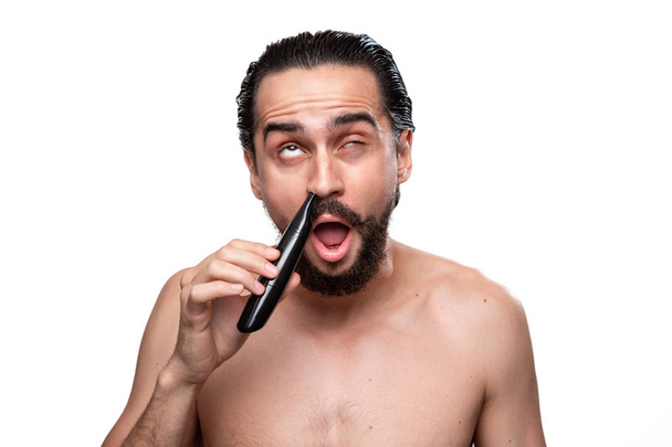 Komea parrakas mies viikset käyttää teho trimmeri leikata hiukset nenässä seisova paljas eristetty valkoisella taustalla. Käsitys aamuhoidosta ja parranajosta
. - Valokuva, kuva