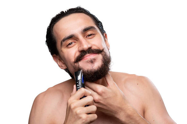 Beau homme avec moustache utilise rasoir électrique pour couper sa barbe debout nu isolé sur fond blanc. Concept de traitement matinal et de rasage. Il est temps de tailler ta barbe. Routine matinale
 - Photo, image
