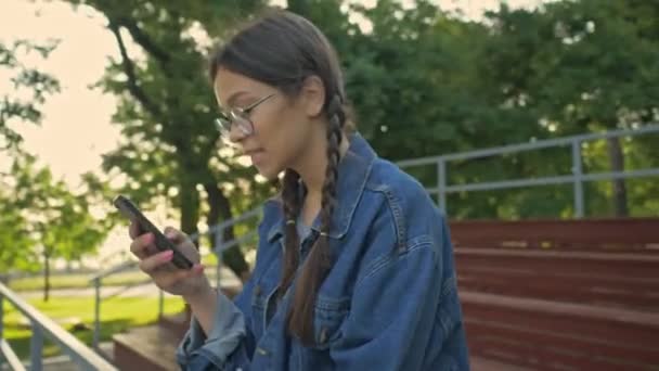 Beautiful excited young brunette girl in denim jacket smiling and chatting on smartphone while sitting in the park - Video, Çekim