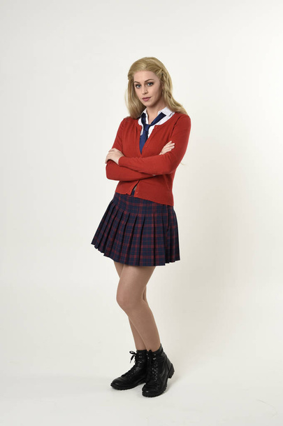 Ganzkörperporträt eines blonden Mädchens in roter Strickjacke mit Krawatte und kariertem Rock, Schuluniform, stehend, in die Kamera blickend, auf weißem Studiohintergrund. - Foto, Bild